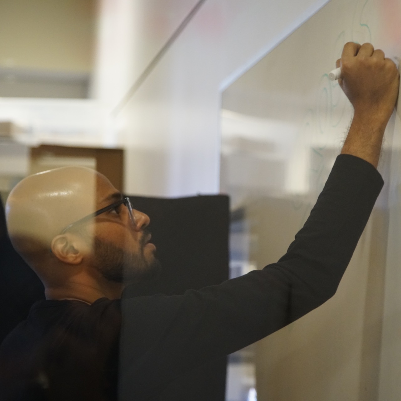 Rishi, seen here at the whiteboard, one of his core natural habitats. A photograph of Rishi drawing at a whiteboard. Rishi is a bespectacled man in his late-twenties, bald with brown skin and a trimmed beard and mustache. He is wearing a black full-sleeved t-shirt, and drawing on a whiteboard in profile.